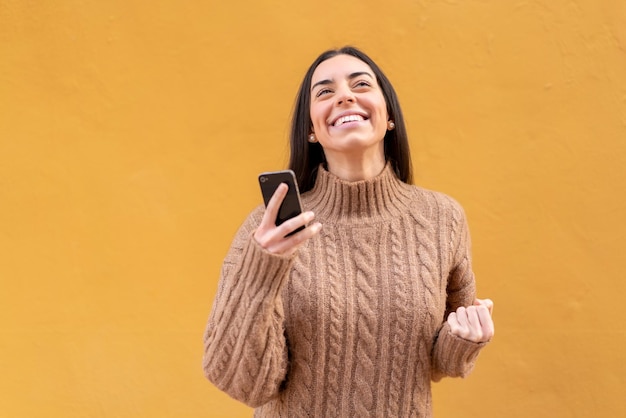 Joven morena al aire libre usando teléfono móvil y haciendo gesto de victoria