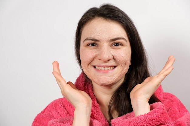 Una joven morena con un albornoz rosa hace que los tratamientos faciales de limpieza sean una máscara o un exfoliante para limpiar y nutrir la piel