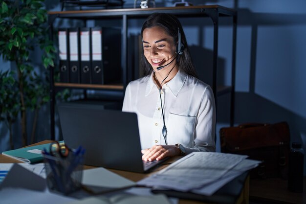 Joven morena con auriculares de agente del centro de llamadas trabajando tarde en la noche con una sonrisa feliz y fresca en la cara. persona con suerte.