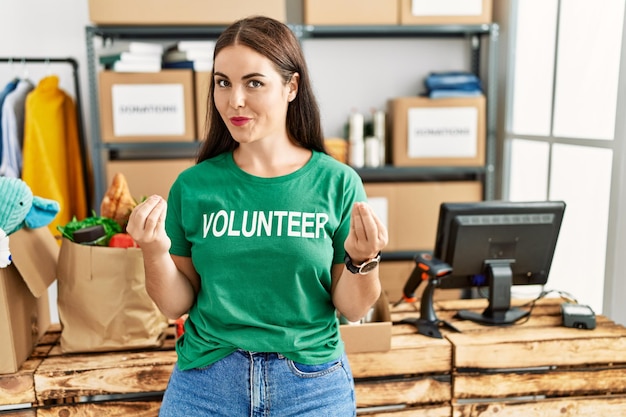 Joven morena con camiseta de voluntaria en el puesto de donaciones haciendo gestos de dinero con las manos pidiendo pago de salario negocio millonario