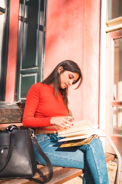 Joven morena caucásica con una camisa roja sentada en un banco en el parque leyendo un libro