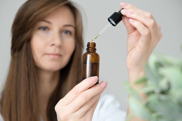 Joven morena caucásica desenfocada haciendo procedimientos de belleza para el cuidado de la piel frente al espejo en casa, sosteniendo una botella de vidrio con suero, aceite, cosméticos bionaturales para el cabello o la cara en casa, de cerca.