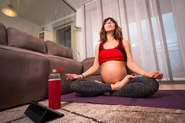 Joven morena caucásica embarazada haciendo ejercicios de relajación de yoga en casa con instrucciones del maestro Ejercicios en línea en clases preparto bienestar durante el embarazo