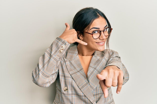 Joven morena con chaqueta de negocios y gafas sonriendo hablando por teléfono y señalándote. llámame.