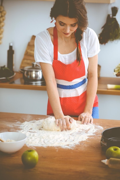 Joven morena cocinando pizza o pasta hecha a mano en la cocina. Ama de casa preparando masa en mesa de madera. Concepto de dieta, alimentación y salud.