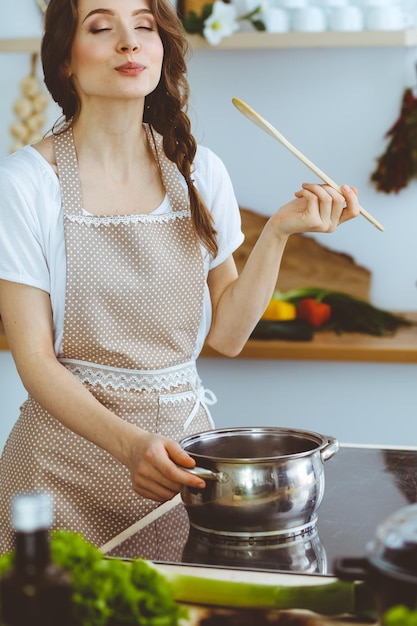 Joven morena cocinando sopa en la cocina. Ama de casa sosteniendo una cuchara de madera en la mano. Concepto de alimentación y salud.