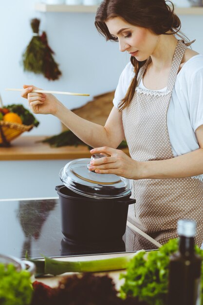 Joven morena cocinando sopa en la cocina. Ama de casa sosteniendo una cuchara de madera en la mano. Concepto de alimentación y salud.