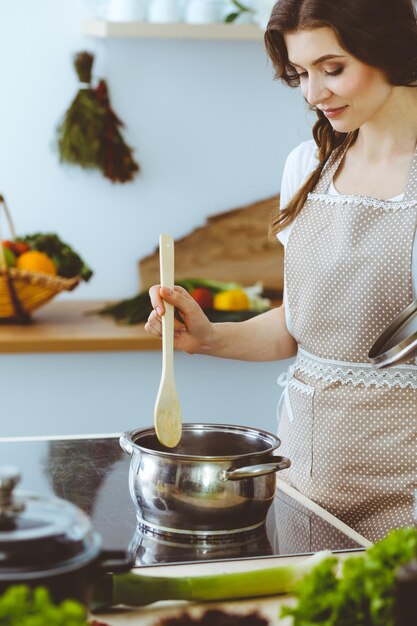 Joven morena cocinando sopa en la cocina. Ama de casa sosteniendo una cuchara de madera en la mano. Concepto de alimentación y salud.