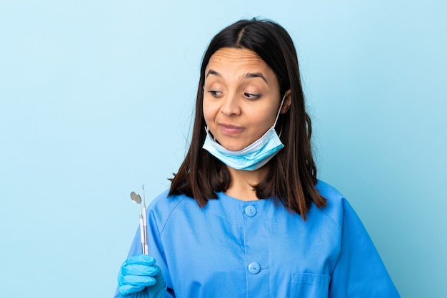 Joven morena dentista de raza mixta mujer sosteniendo herramientas sobre antecedentes aislados teniendo dudas mientras mira de lado