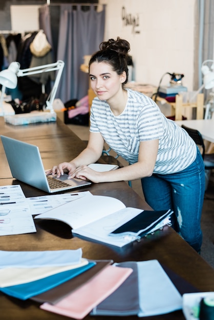 Joven morena diseñadora de moda en ropa casual inclinada sobre la mesa y la computadora portátil mientras se desplaza por las cosas en línea