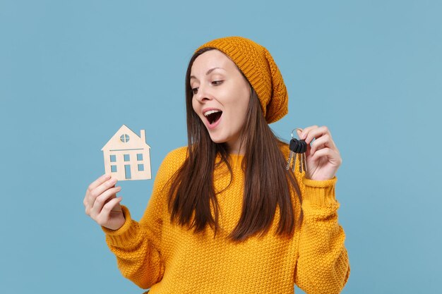 Una joven morena divertida con suéter amarillo y sombrero posando aislada en un retrato de estudio de fondo azul. Gente emociones sinceras concepto de estilo de vida. Simulacros de espacio de copia. Mantenga el manojo de llaves de la casa.