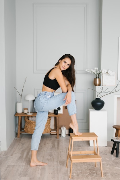 Joven morena elegante con cabello largo en un estilo casual en jeans, un top de punto negro puso su pie en una escalera de tijera en la sala de estar