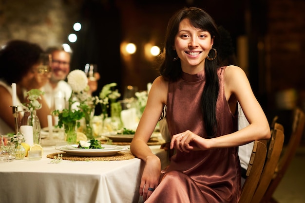 Joven morena elegante con vestido de noche de seda sentada junto a la mesa servida