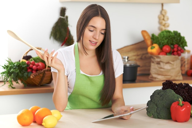 La joven morena está buscando una nueva receta para cocinar en la cocina. Ama de casa sosteniendo una cuchara de madera y una tableta mientras busca en Internet. Concepto de alimentación y salud.