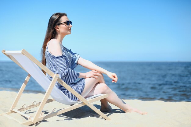 Una joven morena feliz relajándose en una silla de madera en la playa del océano mientras sonríe y lleva gafas de sol de moda.