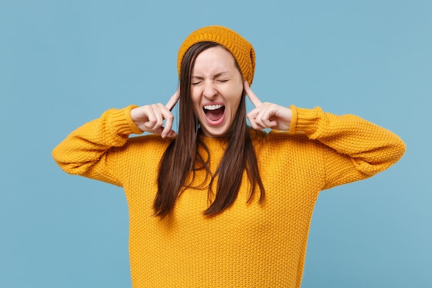 Una joven morena frustrada con suéter amarillo y sombrero posando aislada en un retrato de estudio de fondo azul. Concepto de estilo de vida de las personas. Simulacros de espacio de copia. Gritando, tapándose los oídos con los dedos.