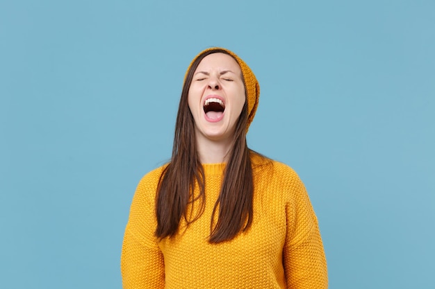 Una joven morena frustrada con suéter amarillo y sombrero posando aislada en un retrato de estudio de fondo de pared azul. Concepto de estilo de vida de las personas. Simulacros de espacio de copia. Gritando, manteniendo los ojos cerrados.