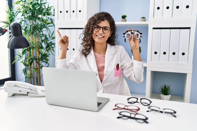 Joven morena con gafas de optometría sonriendo feliz señalando con la mano y el dedo hacia un lado