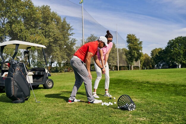 Joven morena con gafas de sol aprendiendo a usar un palo de golf asistida por su entrenador personal