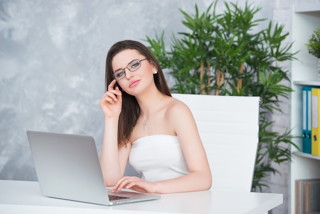 Foto una joven morena con gafas y un vestido blanco está sentada en la mesa de la oficina. la mujer trabaja en la computadora portátil. accesorio de moda o baja visión.