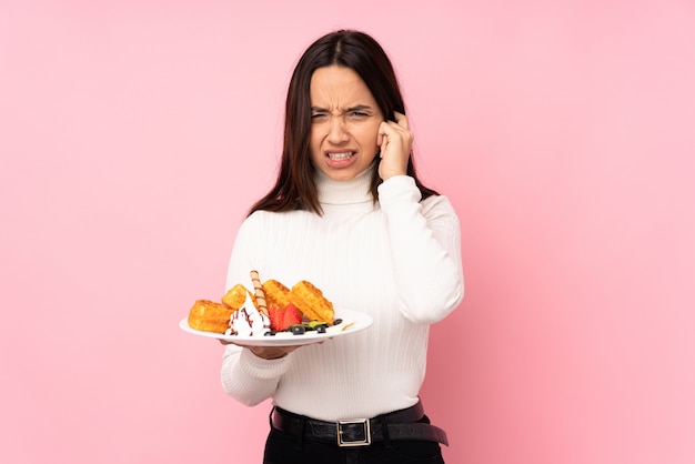 Foto joven morena con gofres sobre pared rosa frustrado y cubriendo las orejas