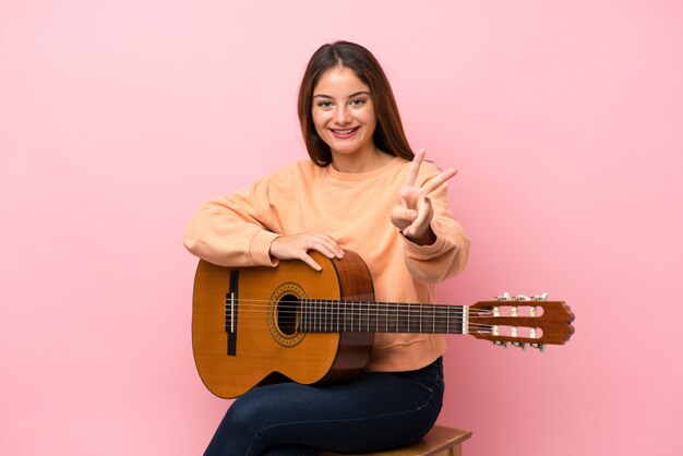 Joven morena con guitarra sobre rosa aislado sonriendo y mostrando el signo de la victoria