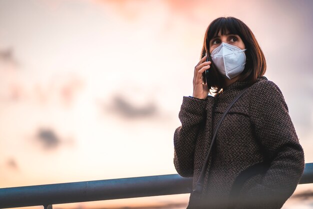 Una joven morena hablando por teléfono con una máscara en una puesta de sol. Primeros pasos de la pandemia incontrolada de Covid-19
