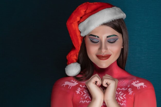 Una joven morena con maquillaje artístico ideal y con un sombrero de Santa Claus pide un deseo con los ojos cerrados