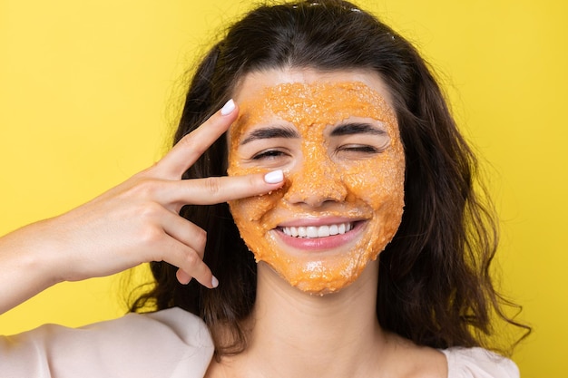 Una joven morena con una mascarilla limpiadora en la cara mira a la cámara y sonríe contra un fondo amarillo