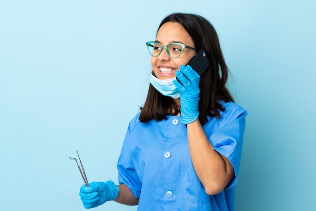 Joven morena mestiza dentista mujer sosteniendo herramientas sobre aisladas manteniendo una conversación con el teléfono móvil