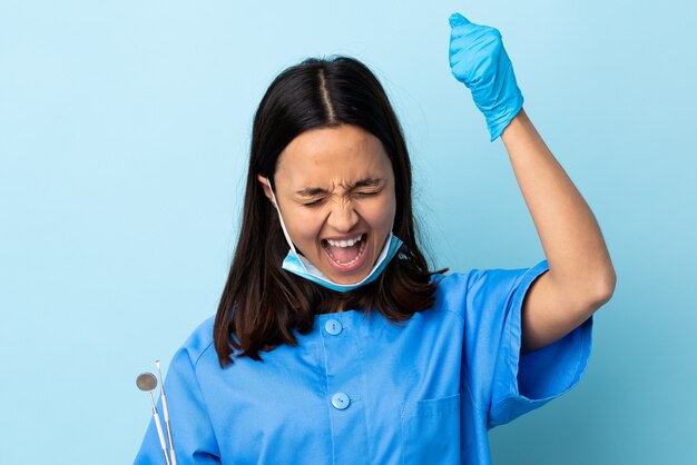 Joven morena mestiza dentista mujer sosteniendo herramientas sobre pared celebrando una victoria