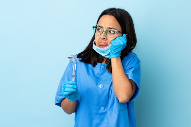 Joven morena mestiza dentista mujer sosteniendo herramientas sobre pared con expresión cansada y aburrida