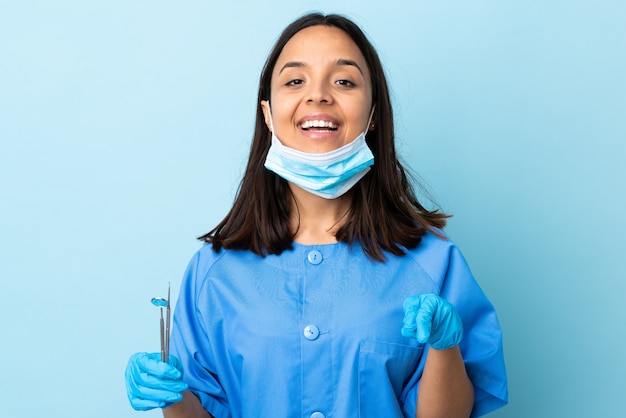 Joven morena mestiza dentista mujer sosteniendo herramientas sobre pared sorprendido y apuntando al frente