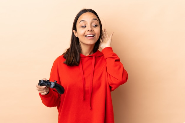 Joven morena mestiza mujer jugando con un controlador de videojuego sobre la pared escuchando algo