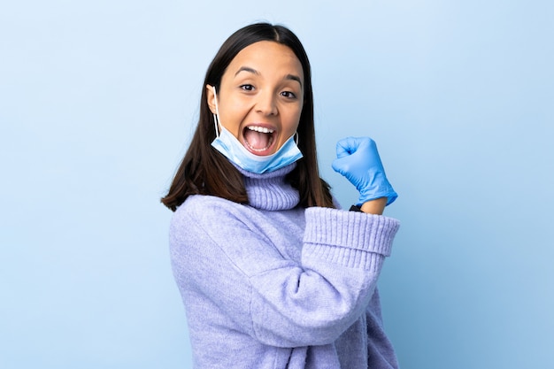 Joven morena mestiza mujer protegiendo con una máscara y guantes sobre pared azul celebrando una victoria