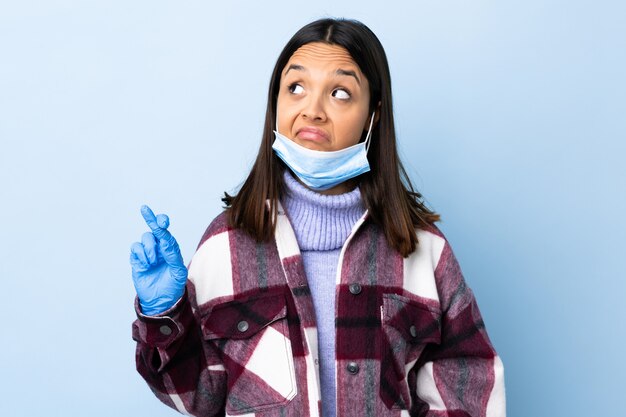 Joven morena mestiza mujer protegiendo con una máscara y guantes sobre la pared azul con los dedos cruzados y deseando lo mejor