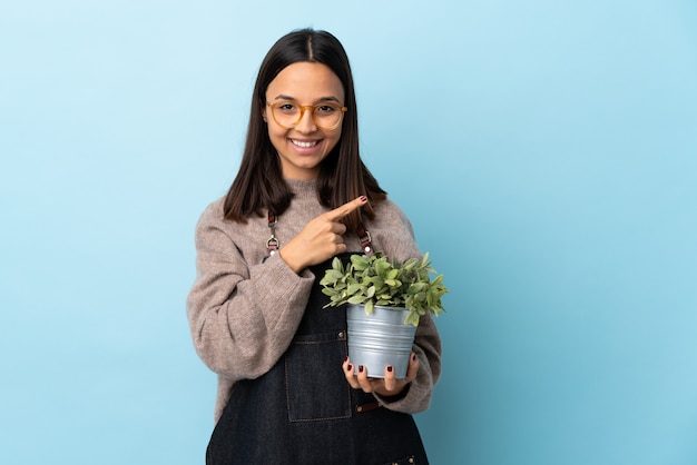 Joven morena mestiza mujer sosteniendo una planta sobre pared azul apuntando hacia un lado para presentar un producto