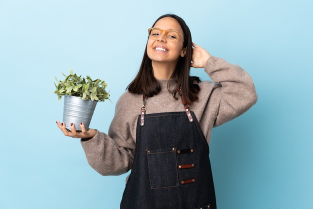 Joven morena mestiza mujer sosteniendo una planta sobre pared azul riendo