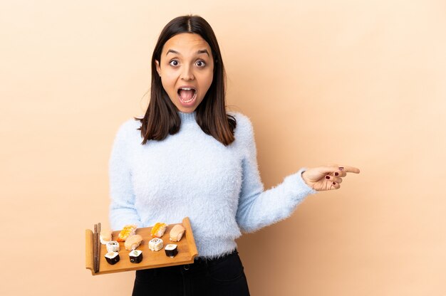 Joven morena mestiza mujer sosteniendo sushi sobre pared aislada sorprendido y apuntando hacia el lado