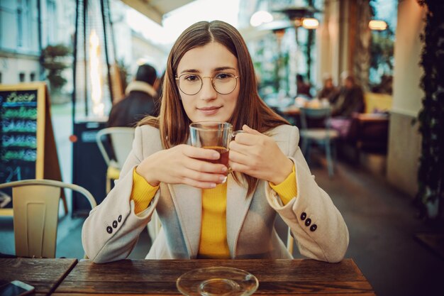 Joven morena de moda sonriente sentado en la terraza de un bar, sosteniendo una taza de té y mirando a la cámara.