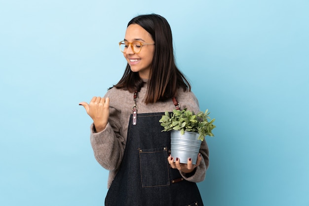 Joven morena mujer de raza mixta sosteniendo una planta sobre pared azul aislada apuntando hacia el lado para presentar un producto.