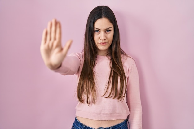 Foto una joven morena parada sobre un fondo rosado deja de cantar con la palma de la mano. expresión de advertencia con gesto negativo y serio en la cara.