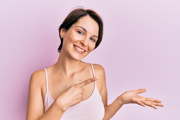 Foto joven morena con el pelo corto sobre fondo rosa asombrada y sonriendo a la cámara mientras presenta con la mano y señala con el dedo