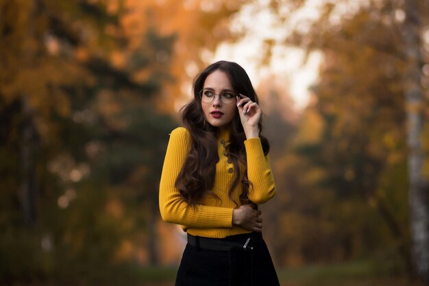 Joven morena de pelo largo con jersey de punto amarillo y anteojos mirando a la cámara mientras se enfrenta al follaje borroso de los árboles de otoño en el parque