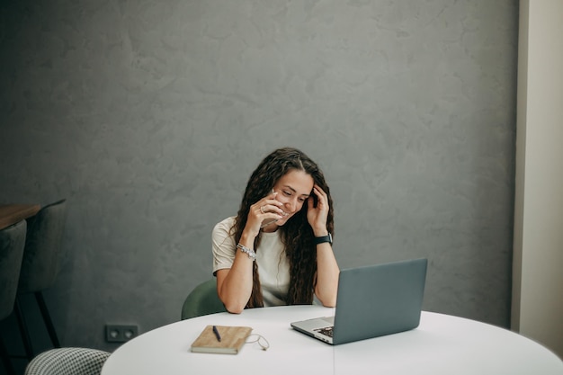 Foto una joven morena con el pelo largo y ondulado en una camiseta beige trabaja en casa en una computadora portátil, habla en un teléfono inteligente y sonríe, un cuaderno con un bolígrafo yace sobre la mesa