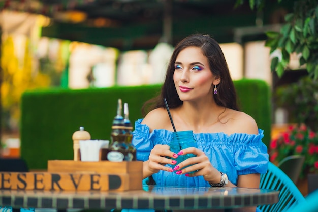 Una joven morena con el pelo largo en la parte superior azul está sentada en la mesa del café y bebiendo de un frasco