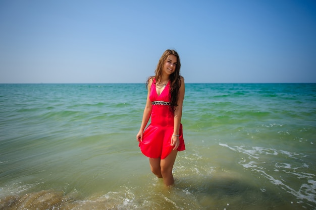 Joven morena de pelo largo sexy en vestido rojo se encuentra en el agua turquesa