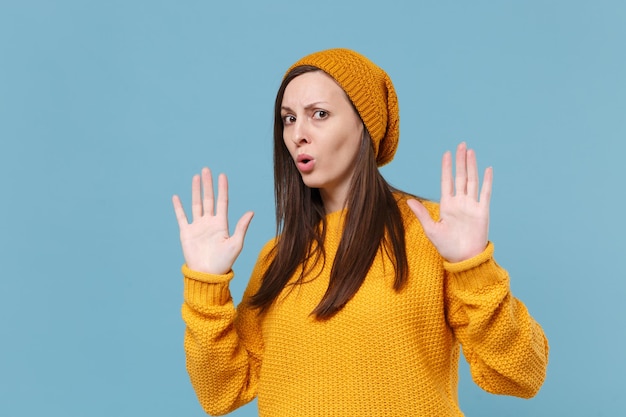 Joven morena perpleja con sombrero de suéter amarillo posando aislada en un retrato de estudio de fondo azul. Gente emociones sinceras concepto de estilo de vida. Simulacros de espacio de copia. Mostrando gesto de parada con las palmas.