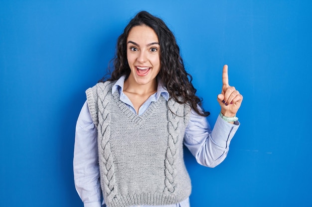 Joven morena de pie sobre fondo azul señalando con el dedo hacia arriba con una idea exitosa, feliz y feliz número uno