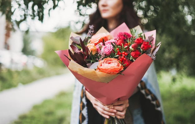 Joven morena con poncho y jeans sostiene un hermoso ramo de flores diferentes en tonos rojos